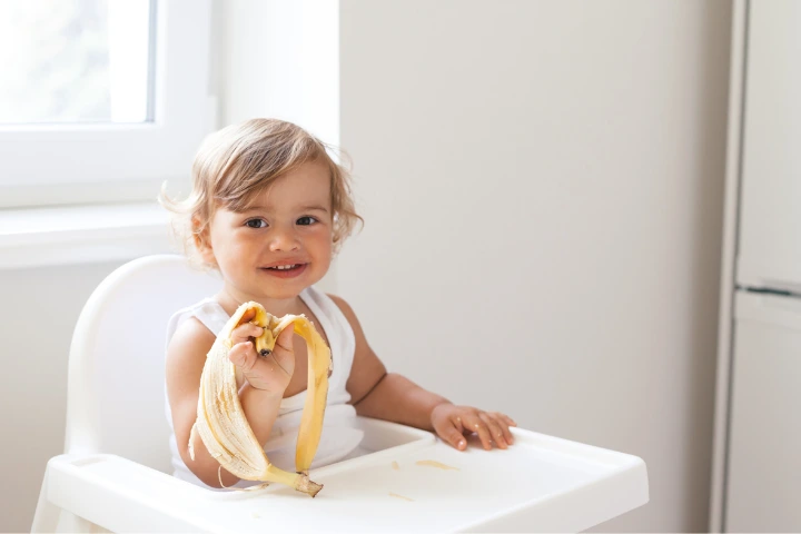 piezas de fruta al dia: bebe comiendo un platano sentado en su sillita